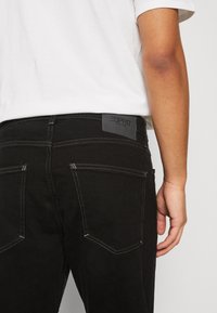 Jeans de mezclilla negra con corte recto, que presentan costuras en contraste, dos bolsillos traseros y un parche de logo en la cintura.