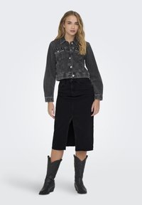 Veste en denim noire raccourcie avec poches avant et fermeture à boutons, associée à une jupe en denim noire avec une fente sur le devant. Portée avec des bottes de cowboy.