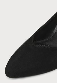 Gros plan sur l'avant et le bord intérieur d'une chaussure plate en daim noir à bout pointu sur un fond uni.