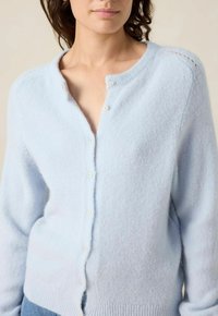 Femme portant un cardigan boutonné en peluche bleu clair avec un col rond et des manches longues, associé à un jean bleu.