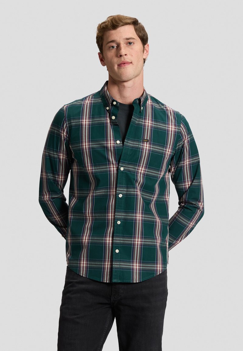Jeune homme portant une chemise à carreaux verte, blanche et rouge à boutons par-dessus un t-shirt noir, debout les mains derrière le dos.