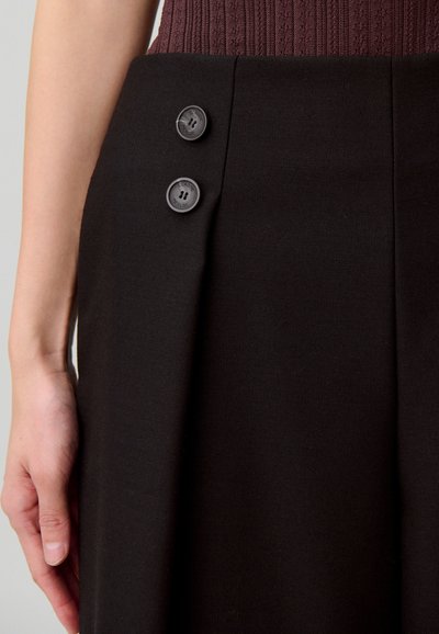 Pantalones negros a medida con una tela suave, dos botones laterales y pliegues nítidos. La cinturilla es de diseño acanalado en color burdeos.