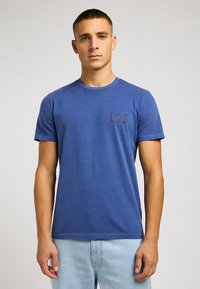 Camiseta de algodón azul con mangas cortas y cuello redondo. Presenta un pequeño logo oscuro en el lado izquierdo del pecho. Textura suave, ajuste casual.