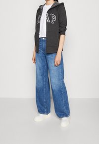 Sweat à capuche gris zippé avec un logo blanc "GAP", porté avec un jean large bleu et des baskets blanches. Apparence décontractée et confortable dans l'ensemble.