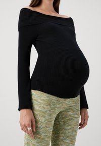 Svart rippad off-shoulder topp med långa ärmar, i kombination med gröna texturerade mönstrade byxor. Modellen visar en tydlig graviditet.