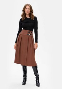 Vrouw in een zwarte top met lange mouwen, een bruine midi rok met hoge taille en gespdetails, en zwarte kniehoge laarzen, staand tegen een witte achtergrond.