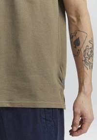Oliven grøn bomulds t-shirt med en struktureret overflade, der har en side split og en lille blå accent. Hånden viser en synlig tatovering.