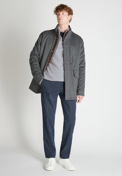 Grau melierte Wolljacke mit braunen Akzenten, mit Reißverschluss am Kragen; darunter ein hellgrauer gerippter Pullover. Dunkelmarineblaue Hose und weiße Turnschuhe.