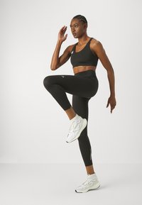 Svart sport-BH og tights med en tettsittende design. Hvite joggesko med teksturerte såler. Modell i en dynamisk posisjon med fletning.