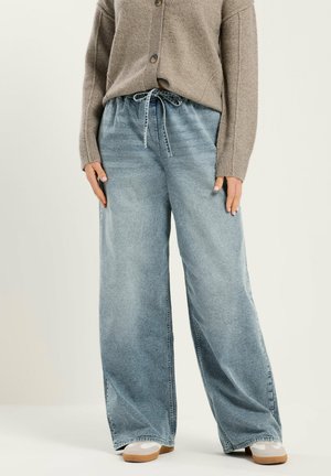 Person in beigem Cardigan, hellblauen Jeans mit weitem Bein und Kordelzug in der Taille sowie weißen Sneakern mit Gummisohlen, die vor einfarbigem Hintergrund steht.