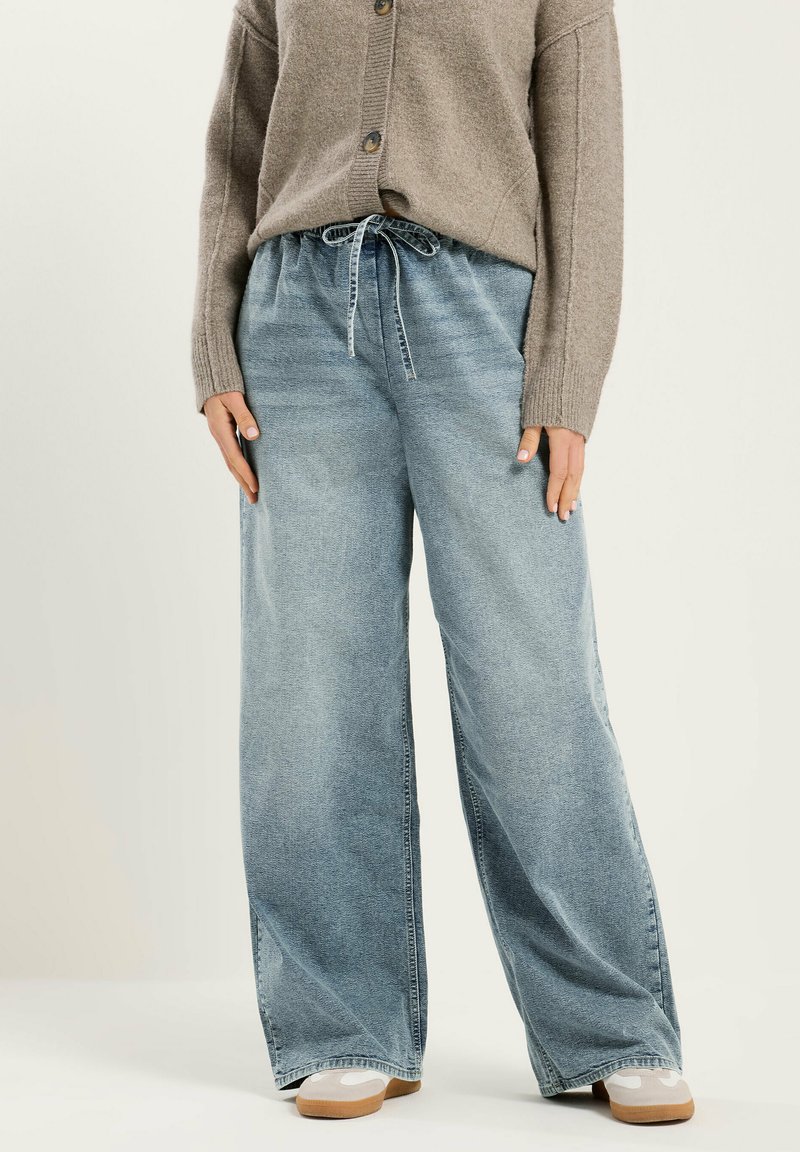 Persona con cárdigan beige, pantalones vaqueros azul claro de pierna ancha con cintura con cordón y zapatillas blancas con suelas de goma, de pie sobre un fondo liso.