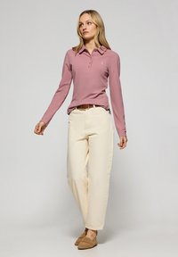 Långärmad, dammig rosa polo med krage och treknapplås, tillsammans med elfenbensfärgade vida byxor och bruna slipskor.