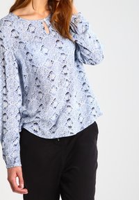 Femme portant une blouse à manches longues bleue à motifs avec une petite ouverture à l'avant et un pantalon noir, posant avec une main près de son visage.