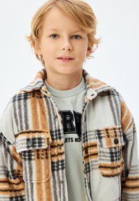 Fleece skjortejakke i hvid, orange og gråternet mønster med to forlommer, trykknapper og en blød tekstur. Let lagdelt over en grøn sweatshirt.