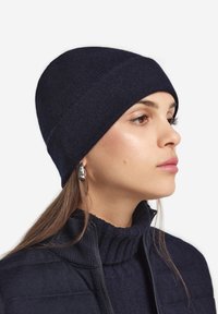 Granatowy dzianinowy czapka beanie o dopasowanym kroju, wykonana z miękkiego materiału, z podwiniętym brzegiem. Modelka nosi pasujący sweter z wysokim kołnierzem oraz srebrne kolczyki.