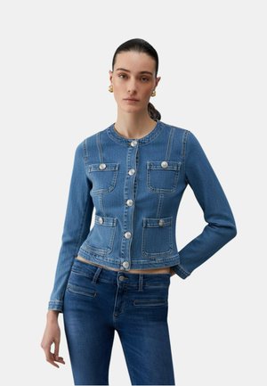 Donna che indossa una giacca di jeans blu aderente con bottoni argentati e jeans blu abbinati, in piedi con una mano sul fianco.