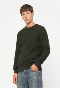 Maglione a maglia verde scuro con un motivo testurizzato, scollatura rotonda e polsini a coste, abbinato a jeans azzurri chiari, mani nelle tasche.