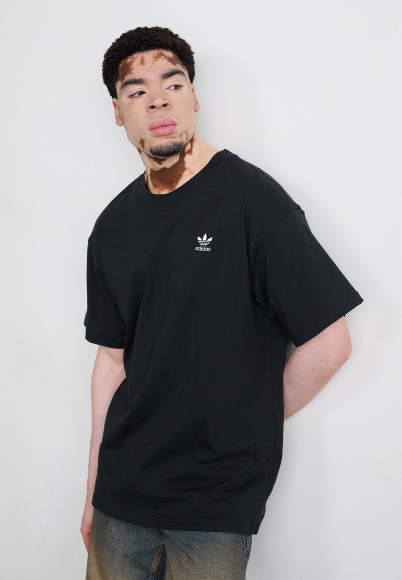 Schwarzes Baumwoll-T-Shirt mit kurzen Ärmeln und Rundhalsausschnitt, das ein kleines weißes Adidas-Logo auf der linken Brust zeigt. Getragen mit hellen Jeans.