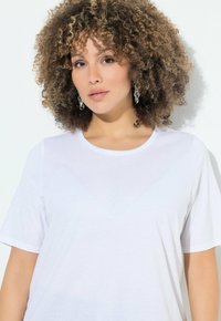 Camiseta blanca de manga corta con cuello redondo. La tela parece suave y lisa. Se lleva con grandes pendientes metálicos.