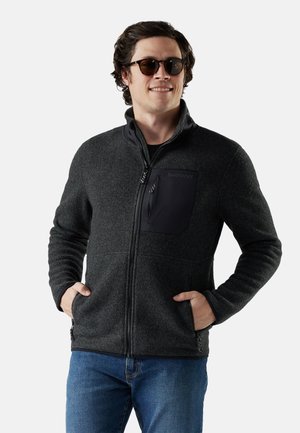 Chaqueta de forro polar gris con un alto cuello, bolsillo en el pecho con cremallera y bolsillos laterales. Cuenta con una textura suave y un diseño ajustado, combinada con jeans azules.