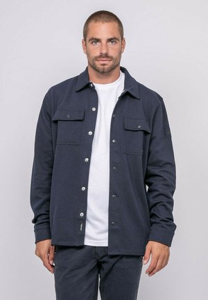 Homme aux cheveux courts et barbe, portant une veste bleu marine par-dessus un t-shirt blanc, se tenant devant un fond gris clair.