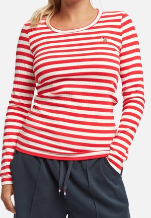 Vrouw draagt een rood-wit gestreept shirt met lange mouwen en een klein geborduurd logo op de borst, en marineblauwe broek met trekkoorden.