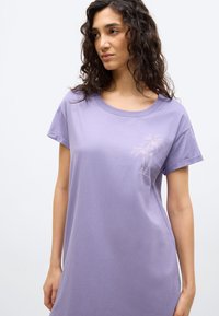 Γυναίκα με μακριά σγουρά μαλλιά, φορώντας φόρεμα T-shirt σε λιλά χρώμα με μικρό σχέδιο φοίνικα στο πάνω μέρος του στήθους, στέκεται μπροστά σε λευκό φόντο.