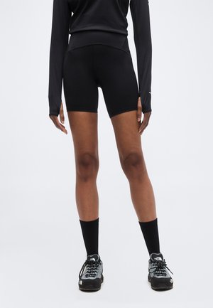 Personne portant un haut à manches longues noir, un short de cyclisme noir, des chaussettes noires et des chaussures de randonnée grises et noires, debout sur un fond clair.