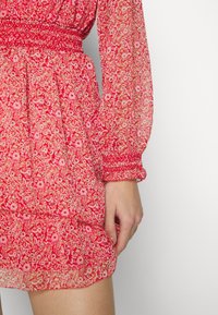 Robe rouge à motif floral avec manches longues transparentes et poignets smockés, portée par une personne avec la main détendue le long du corps.