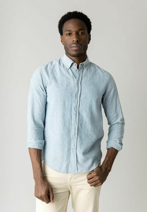 Ung sort mand iført en lyseblå knap-skjorte med op-rullede ærmer og beige bukser, stående mod en ensfarvet grå baggrund.