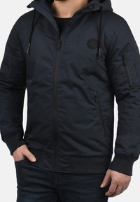 Veste à capuche bleu marine en tissu lisse, dotée d'une fermeture éclair, de poches latérales, de poignets élastiques et d'un patch logo sur la poitrine.