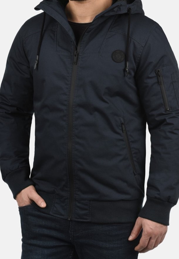 Veste à capuche bleu marine en tissu lisse, dotée d'une fermeture éclair, de poches latérales, de poignets élastiques et d'un patch logo sur la poitrine.