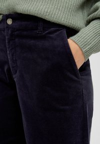Marineblauwe corduroy broek met een gladde textuur, voorzien van zijzakken en een zilveren knoop in de tailleband, gecombineerd met een lichtgroen sweater.