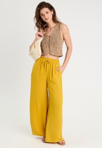 Braunes gestricktes Croptop mit Knopfdetail, kombiniert mit weiten, leuchtend gelben Hosen. Das Model hält eine kleine cremefarbene Tasche und trägt Sandalen.