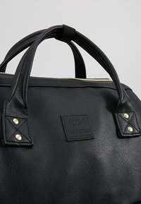 Sac à main noir texturé avec doubles poignées, détails de rivets dorés, et une étiquette de marque rectangulaire cousue à l'avant.