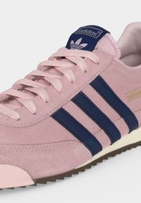 Ροζ σουέντ αθλητικό παπούτσι Adidas με σκούρες μπλε ρίγες, ανοιχτό ροζ κορδόνια και λευκή σόλα με υφή καφέ πάτο.