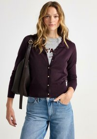 Mørk lilla cardigan med knaplukning, ribbede manchetter og kant, båret over en lys grå skjorte. Stylet med blå jeans og en struktureret sort taske.