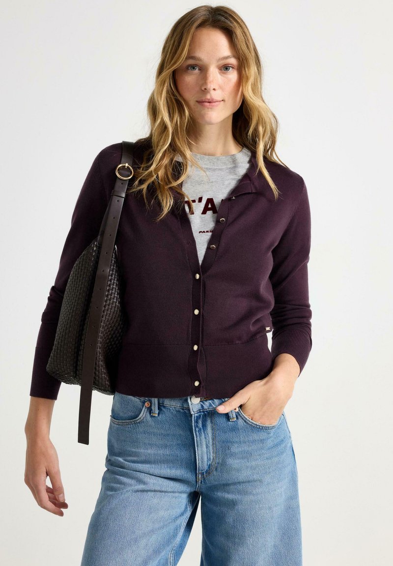 Mørk lilla cardigan med knaplukning, ribbede manchetter og kant, båret over en lys grå skjorte. Stylet med blå jeans og en struktureret sort taske.