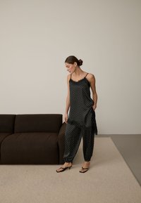 Femme debout à l'intérieur portant un caraco en satin noir à pois et un pantalon ample assorti avec des sandales noires à talons près d'un canapé sombre.
