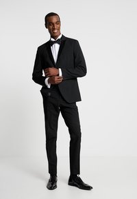 Tuxedo nero con rever satinado, camicia bianca e papillon nero. Pantaloni slim-fit abbinati a scarpe eleganti nere lucide.