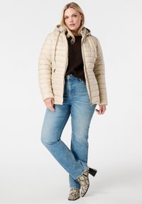 Veste rembourrée beige avec une capuche, dotée d'une fermeture éclair et de poches latérales, portée avec un jean bleu et des bottines à talons imprimé serpent.