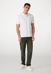 Weißes Polohemd mit Kragen, mit einem kleinen Logo-Patch. Kombiniert mit dunkelgrünen Jeans und weißen Turnschuhen. Glattes Gewebe, lässige Passform.
