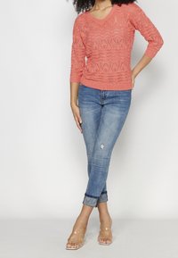 Pull en maille corail avec col en V, présentant un motif texturé ; associé à un jean denim bleu clair déchiré et des sandales à talons blocs transparents.