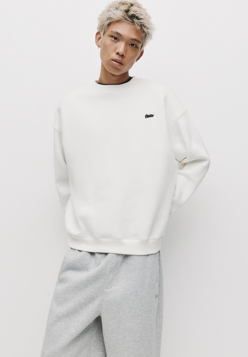Sweat-shirt blanc avec des poignets et un ourlet côtelés, arborant un petit logo noir sur la poitrine. Porté avec un pantalon de survêtement gris clair, ample.