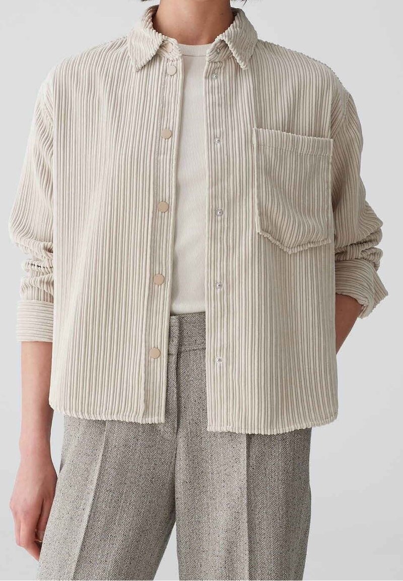 Chemise boutonnée en velours côtelé beige avec un col, une poche avant et une texture côtelée, portée par-dessus un haut ajusté blanc et un pantalon gris à motifs.