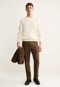 Maglione a maglia beige chiaro e pantaloni marroni su misura, abbinati a scarpe scure. L'individuo tiene un blazer marrone sotto il braccio.