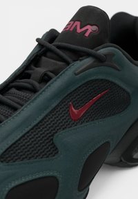 Zatvoren kadar crne i tamnozelene tenisice s mrežastim detaljima, crnim vezicama i crvenim Nikeovim logotipom sa strane.
