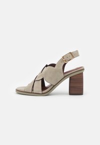 Beige suede sandal with a slotted design, open toe, and slingback strap featuring a gold buckle. Block heel with a wood-like finish.