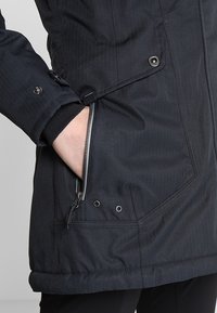 Main dans la poche d'une veste noire zippée avec bouton-pression et aérations, montrant le poignet de la manche avec fermeture à bouton-pression.