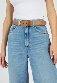Brun, texturerat bälte med silverfärgad metallsp�änne bärs över ljusblå, högmidjade jeans. Jeansens tyg har en lätt urtvättning och en lös passform.